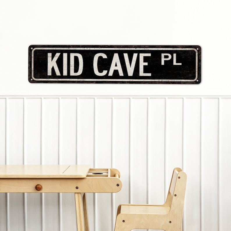 May include: A black and white sign with the text "KID CAVE PL" on a white wall. A light wood desk and chair are in the foreground.