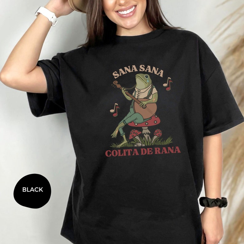 K&ouml;nnte beinhalten: Schwarzes Oversize-T-Shirt mit einem gr&uuml;nen Frosch, der auf einem Pilz Banjo spielt. Der Text "Sana Sana Colita De Rana" ist auf dem Shirt aufgedruckt.