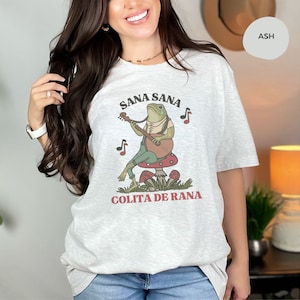 K&ouml;nnte beinhalten: Ein hellgraues T-Shirt mit einem gr&uuml;nen Frosch, der auf einem roten Pilz Gitarre spielt. Der Text "Sana Sana Colita de Rana" ist auf dem T-Shirt aufgedruckt.