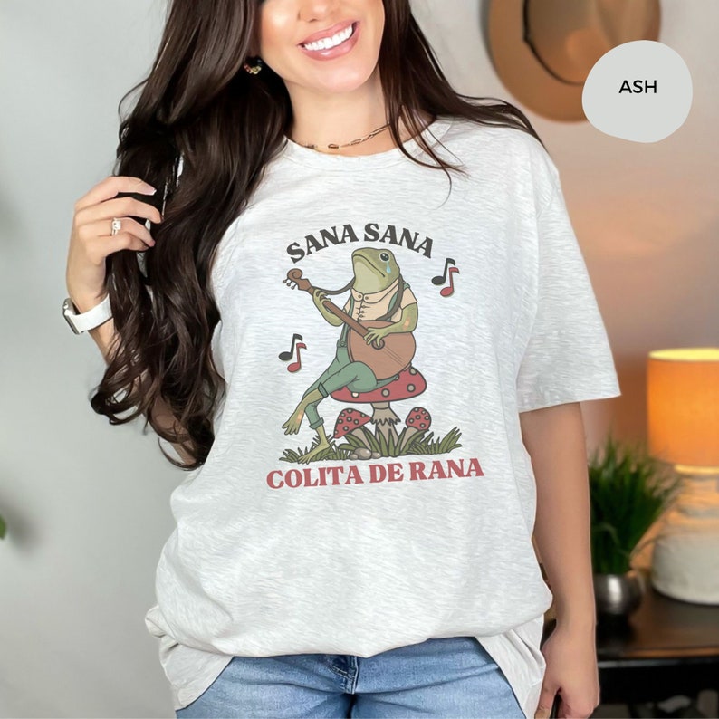 K&ouml;nnte beinhalten: Ein hellgraues T-Shirt mit einem gr&uuml;nen Frosch, der auf einem roten Pilz Gitarre spielt. Der Text "Sana Sana Colita de Rana" ist auf dem T-Shirt aufgedruckt.