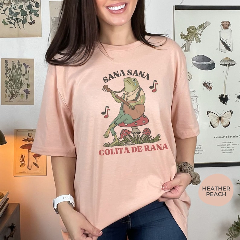 K&ouml;nnte beinhalten: Ein hellrosa T-Shirt mit einem Cartoon-Frosch, der Gitarre spielt, und dem Text "Sana Sana Colita de Rana" auf der Vorderseite.