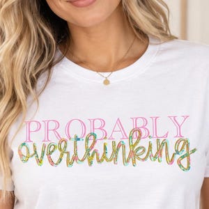 May include: A white short-sleeved t-shirt featuring the text "PROBABLY" in pink capital letters and the word "overthinking" in a cursive, multi-colored floral pattern. The shirt is paired with light blue denim jeans with a visible metal button.