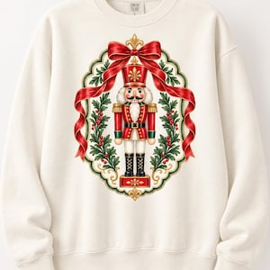 May include: A cream-colored crewneck sweatshirt featuring a central graphic of a nutcracker soldier in a red and gold uniform. The nutcracker is framed by a decorative oval border of green holly leaves, red berries, and a large red ribbon bow.