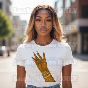 May include: White t-shirt featuring a graphic of a hand with long, pink-painted nails and gold bracelets. The text "Lucky Vibes Only" is printed on the shirt. The model is wearing blue jeans.