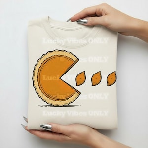 Op de afbeelding: Crèmekleurig T-shirt met een taartpuntontwerp in de stijl van Pac-Man, met drie taartstukjes. De taart is oranje met een bruine korst. De tekst "Lucky Vibes ONLY" staat op het shirt.