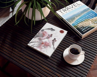 May include: A white book with a watercolor painting of koi fish and a red sun on a dark wood table. The book is next to a magazine titled "Travel Magazine" with a photo of a beach and text in a foreign language. A white cup of coffee sits on a white saucer on the table.