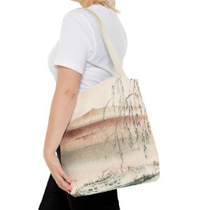 Puede incluir: Una bolsa de tela beige con un diseño de acuarela de un paisaje con un río y sauces. El diseño está en tonos de marrón, verde y blanco.