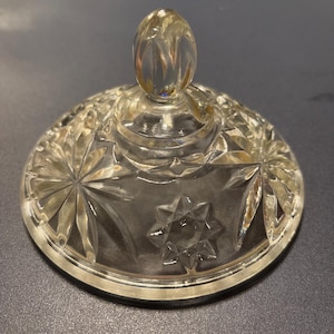 May include: A clear, glass lid with a decorative starburst and star pattern. The lid has a rounded top with a textured, oval-shaped handle. The glass is transparent, allowing light to pass through, and is likely for a jar or container.