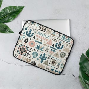 May include: A laptop sleeve with a cream background and a repeating pattern of cacti, triangles, and geometric shapes in teal, pink, and brown. The sleeve has a black zipper and trim. The design has a Southwestern or bohemian theme.