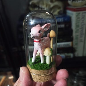May include: A pink deer figurine with silver glitter accents and a red collar is displayed inside a glass cloche. The miniature scene includes artificial mushrooms and green moss on a cork base, creating a whimsical, decorative item.