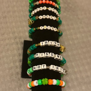 May include: A collection of beaded bracelets in green, white, and orange, with text spelling out "LUCKY", "IRISH", "KISS ME", and "NOT IRISH". Some bracelets feature shamrock charms. The bracelets are displayed on a black stand.