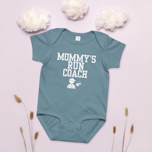 Op de afbeelding: Een turquoise baby-romper met de witte tekst "MOMMY'S RUN COACH". Daaronder een witte afbeelding van een persoon met een hoed en stopwatch. De achtergrond toont pluizige wolken en gedroogde planten, wat een zachte uitstraling creëert.