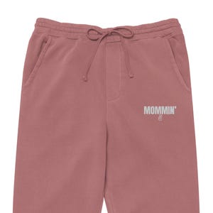 May include: Dusty rose-coloured sweatpants with a drawstring waist and pockets. The text "MOMMIN'" is printed in silver on the left leg, along with a peace sign. These comfortable pants are perfect for casual wear.