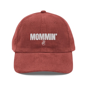 May include: A corduroy baseball cap in a reddish-brown hue. The cap features the word "MOMMIN'" embroidered in white, with a peace sign below. The hat has a curved brim and an adjustable strap at the back.