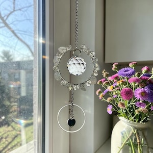 May include: A hanging suncatcher featuring a silver-toned wire hoop adorned with clear, irregular crystal beads. A faceted clear glass sphere hangs in the center, while a smaller silver hoop with a tiny black heart charm dangles below.