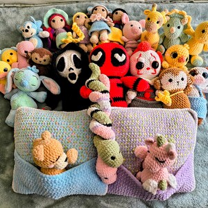 May include: A collection of various crocheted plush toys including characters like Deadpool, Ghostface, and a clown, alongside animals like a giraffe, monkey, and duck. A large rectangular pillow with two pockets holds a small bear and a unicorn plush.