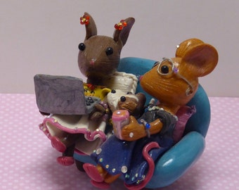 May include: A whimsical figurine featuring a brown bunny and a tan mouse seated on a blue chair. The bunny is using a laptop, while the mouse holds a pink cup. Both are adorned with decorative clothing and accessories. The figurine is set against a pink polka dot background.