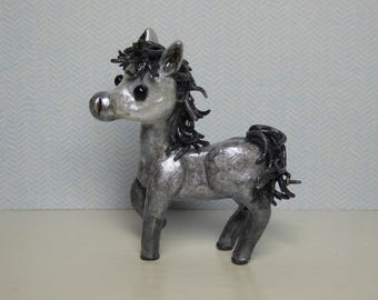 May include: A small, gray, silver-painted horse figurine with black yarn hair and tail. The horse has a whimsical expression and is standing on a white surface.