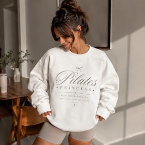 Può includere: Una felpa color crema Pilates Princess con girocollo e maniche lunghe. La parte anteriore presenta le parole "Pilates Princess" in un carattere corsivo, con le parole "Girlhood Society" e altro testo sottostante. La felpa è indossata da una persona.