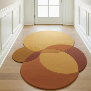 May include: A multi-colored, circular area rug with overlapping sections in shades of gold, brown, and rust. The rug is placed on a hardwood floor in a hallway, with a white door and trim in the background. The rug's design is geometric and modern.