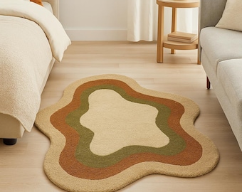 May include: A beige, green, and brown wavy-shaped rug on a hardwood floor. The rug has a layered design with a light beige center, surrounded by green and brown bands. The rug is placed in a room with a bed, sofa, and side table.