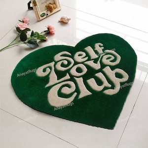 May include: A heart-shaped green rug with the words "Self Love Club" in white, stylized lettering. The rug is placed on a white tiled floor. The brand name "AnayatRugs" is printed on the rug.