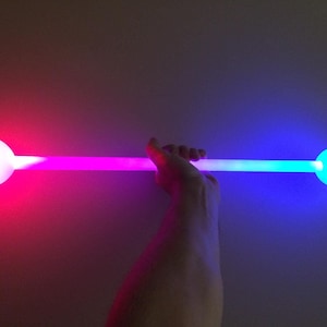 May include: A glowing light stick held horizontally, with a white sphere at each end. One end glows red, the other blue, with a purple gradient in between. The background is a neutral colour.