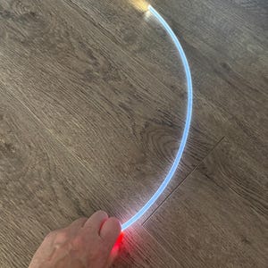 May include: A curved, glowing fiber optic cable with a gradient of colors. The cable starts with a red glow and transitions to blue, ending with a yellow-white light. The cable is lying on a wooden floor.