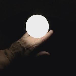 Può includere: Una sfera bianca brillante tenuta in una mano aperta su uno sfondo nero. L'orbita emette una luce soffusa e diffusa, creando un forte contrasto con l'oscurità. La mano è parzialmente visibile, con l'attenzione sulla sfera illuminata.