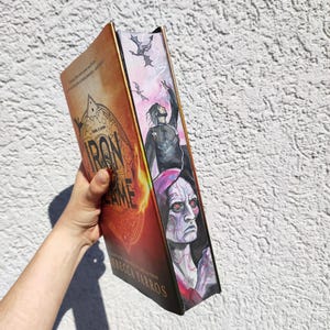 Op de afbeelding: Een paperback boek getiteld "Iron in the Game" van Rebecca Yarros. De omslag heeft een vurige oranje en rode achtergrond met een cirkelvormig ontwerp en de titel in zwart. De rug heeft een kleurrijke illustratie van een persoon met een opgeheven hand en een draak die erboven vliegt.