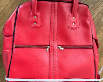 May include: A red bowling bag with black trim and two zippered pockets. The bag has two handles and measures approximately 12 inches across. The bag has a textured surface and a vertical stitched seam down the center.