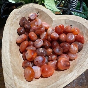 May include: A wooden heart-shaped bowl filled with carnelian tumbled stones. The stones are a mix of orange, red, and white.