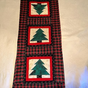 May include: A Christmas-themed table runner featuring a red and black plaid pattern. The runner has four square appliqués with green Christmas tree designs on a cream background, framed in red and black.
