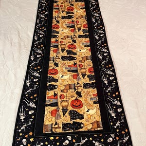 May include: A rectangular fabric table runner featuring a central panel with a yellow background printed with pumpkins, black cats, and crescent moons, bordered by a black fabric frame with a repeating pattern of white skeletons and orange leaves.