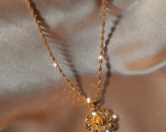 May include: A gold-colored necklace with a flower pendant. The pendant has a detailed floral design with multiple layers of petals. The chain is a twisted rope style. The necklace is displayed on a satin surface.
