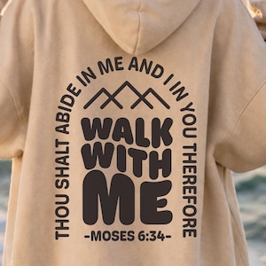 May include: A beige hooded sweatshirt featuring a dark brown graphic on the back. The design includes a mountain icon above the text "WALK WITH ME" and the surrounding phrase "THOU SHALT ABIDE IN ME AND I IN YOU THEREFORE -MOSES 6:34-".