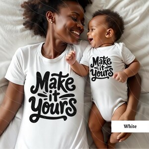 May include: A white t-shirt with the text "Make it yours" in black lettering. A matching white bodysuit with the same text is worn by a baby. The baby is being held by an adult wearing the t-shirt.