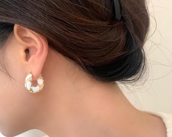 May include: Gold-tone hoop earrings with a white enamel inlay. The earrings have a chunky design with a woven pattern. The earrings are a stylish accessory for everyday wear.