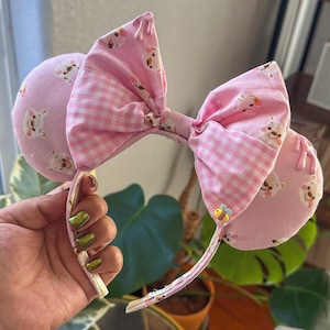 May include: Pink and white fabric headband with large ears and a bow. The ears are round and covered in a light pink fabric with a repeating bear pattern. The bow is a gingham pattern with a bear pattern. A small bee charm is attached.