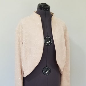 May include: A light peach-colored bolero jacket with long sleeves. The jacket is embellished with small, shimmering beads arranged in a scalloped pattern. The jacket is displayed on a dark gray dress form against a plain white background.