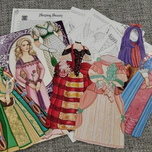 K&ouml;nnte beinhalten: Eine Sammlung von Papierpuppen und zugeh&ouml;rigen Materialien, darunter Illustrationen historischer Kleider und einer Prinzessin. Die Puppen sind in verschiedenen Farben gehalten, darunter Rot, Gr&uuml;n, Rosa und Blau. Der Text "Sleeping Beauty" ist sichtbar.