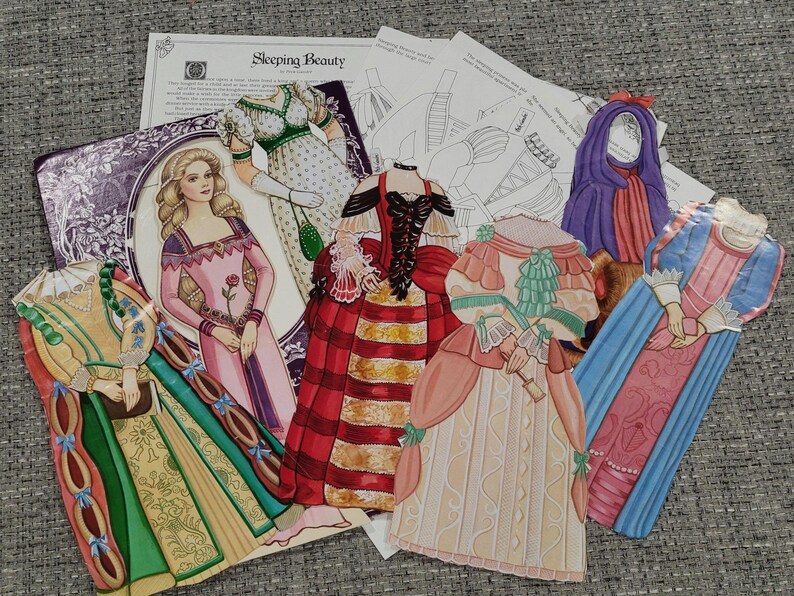 K&ouml;nnte beinhalten: Eine Sammlung von Papierpuppen und zugeh&ouml;rigen Materialien, darunter Illustrationen historischer Kleider und einer Prinzessin. Die Puppen sind in verschiedenen Farben gehalten, darunter Rot, Gr&uuml;n, Rosa und Blau. Der Text "Sleeping Beauty" ist sichtbar.