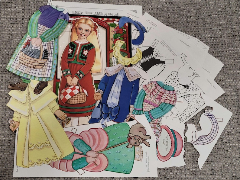 K&ouml;nnte beinhalten: Eine Sammlung von Vintage-Papierpuppen und -Outfits mit Illustrationen aus "Rotk&auml;ppchen". Das Set enth&auml;lt ein M&auml;dchen in einem roten Kleid, eine Frau in einem gr&uuml;nen und lila Kleid und verschiedene Kleidungsst&uuml;cke in Gelb, Blau und Rosa. Einige Teile sind ausgeschnitten.