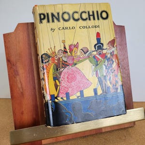 May include: A vintage copy of "Pinocchio" by Carlo Collodi, displayed in a wooden book stand. The book cover features a colourful illustration of characters from the story. The stand has a brass-coloured metal support.