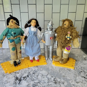 May include: Four dolls from the Wizard of Oz: Scarecrow, Dorothy, Tin Man, and Cowardly Lion. The Scarecrow wears a blue jacket and patched pants. Dorothy has a blue gingham dress. The Tin Man is silver. The Lion has a brown mane.
