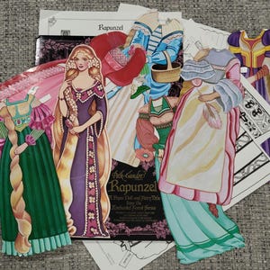 K&ouml;nnte beinhalten: Eine Sammlung von Vintage-Papierpuppen und Outfits mit dem Thema Rapunzel. Die Puppen sind in farbenfrohen Kleidern und Gew&auml;ndern mit langen, flie&szlig;enden Haaren dargestellt. Das Set enth&auml;lt eine Puppe, Outfits und ein Heft.