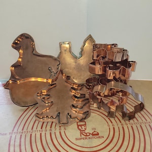 Puede incluir: Un conjunto de cortadores de galletas de color cobre en varias formas, incluyendo un cordero, un árbol de Navidad, una estrella y un hombre de jengibre. Los cortadores están sobre una esterilla de silicona roja y blanca.