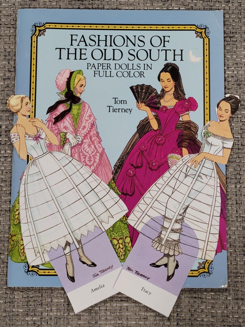 K&ouml;nnte beinhalten: Ein Vintage-Papierpuppenbuch mit dem Titel "Fashions of the Old South" mit Illustrationen von Frauen in historischen Kleidern. Das Cover zeigt vier Frauen in farbenfrohen Kleidern, mit zwei Papierpuppen mit der Aufschrift "Amelia" und "Tracy".