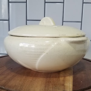 Puede incluir: Una cacerola de cerámica blanca con tapa. La cacerola tiene un borde festoneado y un diseño floral. La tapa tiene un pomo en la parte superior.