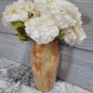 May include: A decorative vase with a swirling, marbled pattern in shades of brown and tan. The vase holds a bouquet of large, cream-colored artificial flowers with green leaves. The arrangement is set against a gray wooden background.